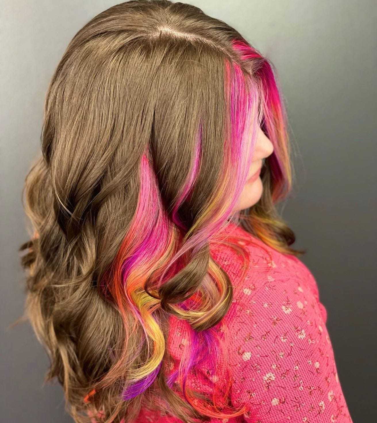 Woman with long brown hair highlighted with vibrant pink, yellow, and orange streaks, wearing a pink shirt.