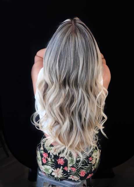 Woman with long, wavy blonde hair sitting, wearing floral-patterned shorts against a dark background.