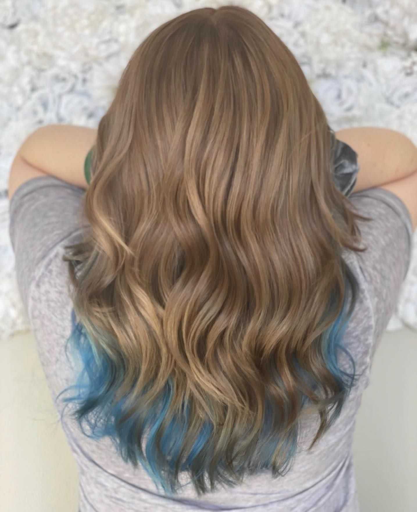 Woman with long wavy brown hair and blue-dyed tips, wearing a gray shirt, seen from behind.