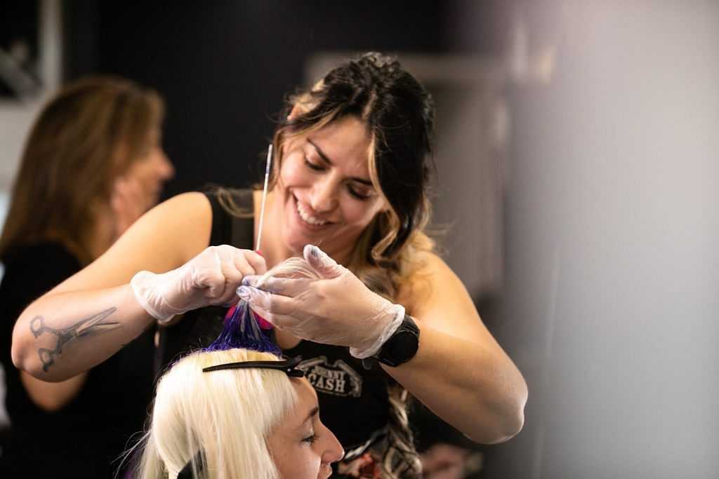 Hairdresser dyeing client's hair purple while smiling, another woman in background.
