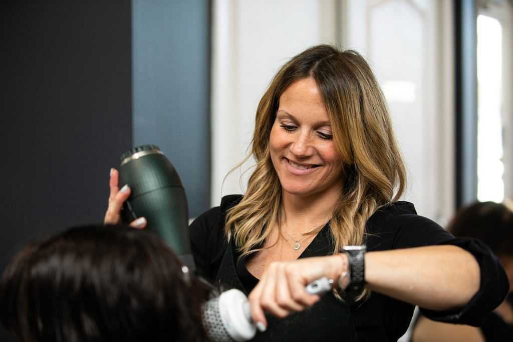 Woman smiling while blow-drying a client's hair in a salon.
