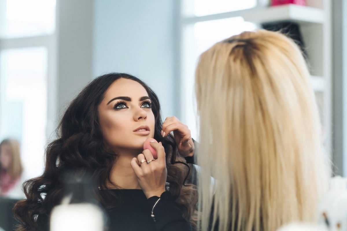 A makeup artist applying foundation to a woman with long, dark, wavy hair in a bright room.