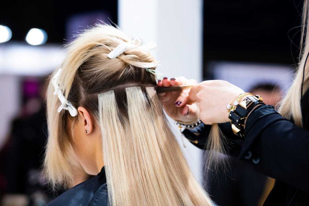 Hair stylist applying extensions to a client's blonde hair at a salon.